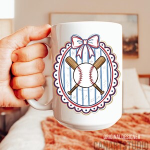 May include: White ceramic mug featuring a baseball design. The design includes a baseball, crossed bats, and a bow, all within a decorative oval frame. The mug has the text "ORIGINAL DESIGNER CANARTSTUDIO" at the bottom.