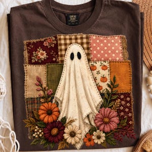 Puede incluir: Una camiseta marrón oscuro con un gráfico central de un fantasma blanco rodeado por un diseño de patchwork. Los cuadrados del patchwork incluyen patrones florales, de cuadros y de lunares, acentuados con flores bordadas naranjas y rosas.