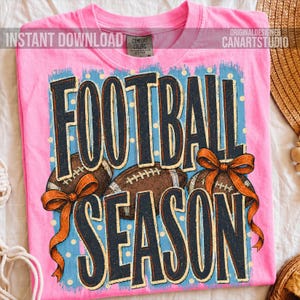 K&ouml;nnte beinhalten: Ein leuchtend pinkes T-Shirt mit dem Schriftzug FOOTBALL SEASON in gro&szlig;en dunkelblauen Buchstaben. Das Design zeigt zwei braune Footballs und zwei orangefarbene Schleifen auf hellblauem Grund mit wei&szlig;en Punkten. Das Shirt hat ein Comfort Colors Etikett.