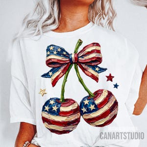 Puede incluir: Camiseta blanca con un diseño patriótico de dos cerezas con un motivo de la bandera estadounidense, un lazo y estrellas. Las cerezas son rojas, blancas y azules, con un tallo verde. El texto "CANARTSTUDIO" es visible.