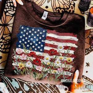 May include: A brown sweatshirt featuring a painted American flag design with red and white stripes, blue stars, and colorful flowers. The text "ORIGINAL DESIGN STUDIO" is visible.