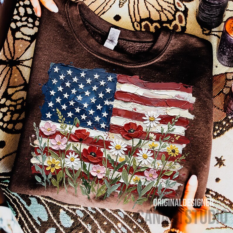 May include: A brown sweatshirt featuring a painted American flag design with red and white stripes, blue stars, and colorful flowers. The text "ORIGINAL DESIGN STUDIO" is visible.