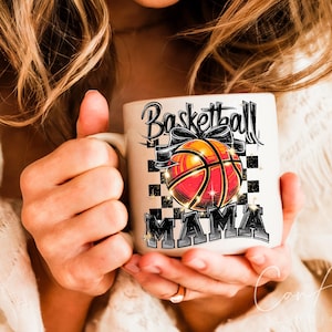 May include: White ceramic mug with a black and white chequered background. The mug has a black and white graphic of a basketball with a bow and the text "Basketball Mama".