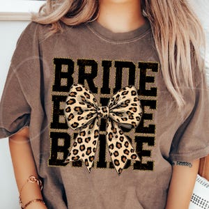 May include: A brown t-shirt with the word "BRIDE" printed in black with gold glitter accents. A large leopard print bow is centred on the shirt.