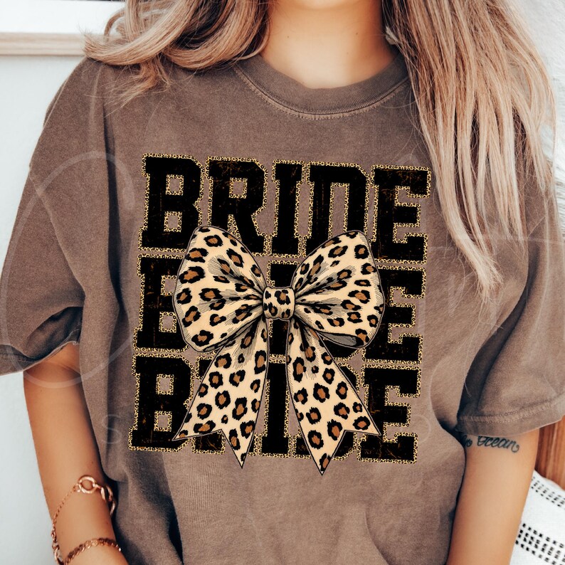 May include: A brown t-shirt with the word "BRIDE" printed in black with gold glitter accents. A large leopard print bow is centred on the shirt.