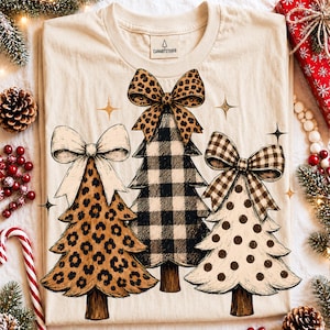 May include: A cream-colored t-shirt featuring a graphic of three stylized Christmas trees. The left tree has a leopard print with a cream bow, the center tree has a black and white buffalo plaid pattern with a leopard print bow, and the right tree has brown polka dots with a brown and white gingham bow.
