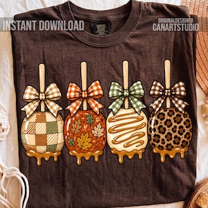 May include: A dark brown t-shirt featuring a graphic of four caramel apples on sticks, each topped with a patterned bow. The apples display designs including green and brown plaid, autumn leaves, caramel drizzle, and a brown leopard print pattern.