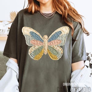 May include: Dark gray t-shirt featuring a large, colorful butterfly applique. The butterfly design incorporates various fabric patterns, including gingham and floral prints, in shades of blue, pink, and yellow. The text "ORIGINAL DESIGNER CANA STUDIO" is printed on the bottom right.