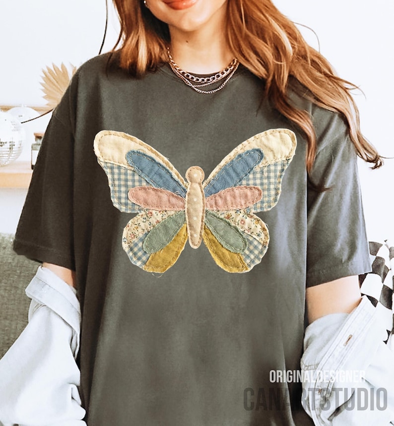 May include: Dark gray t-shirt featuring a large, colorful butterfly applique. The butterfly design incorporates various fabric patterns, including gingham and floral prints, in shades of blue, pink, and yellow. The text "ORIGINAL DESIGNER CANA STUDIO" is printed on the bottom right.