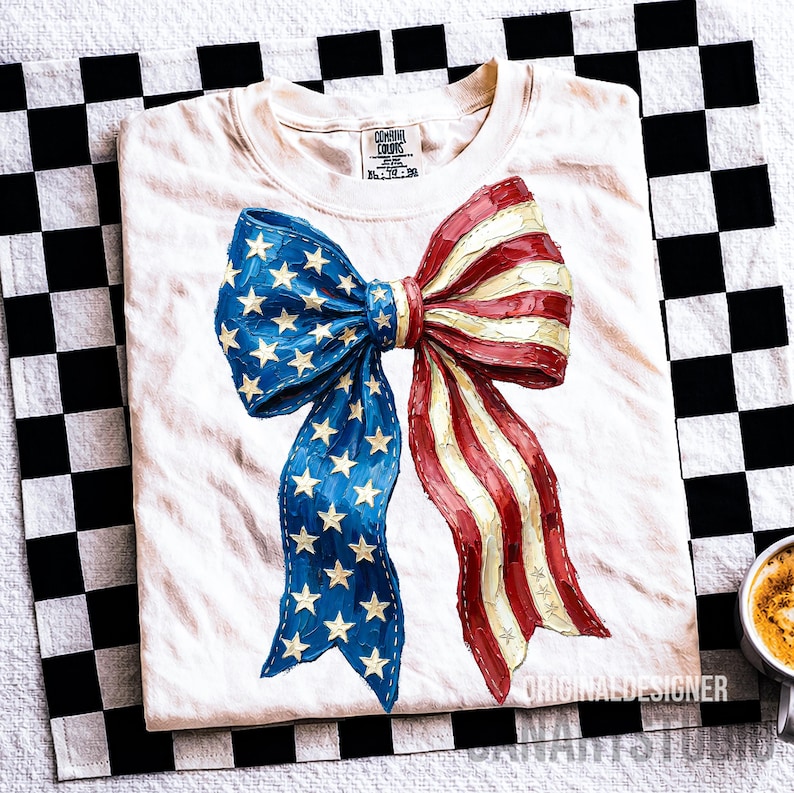 May include: Off-white t-shirt with a large bow design resembling the American flag. The bow features blue sections with white stars and red and white stripes. The shirt is folded on a black and white chequered surface.