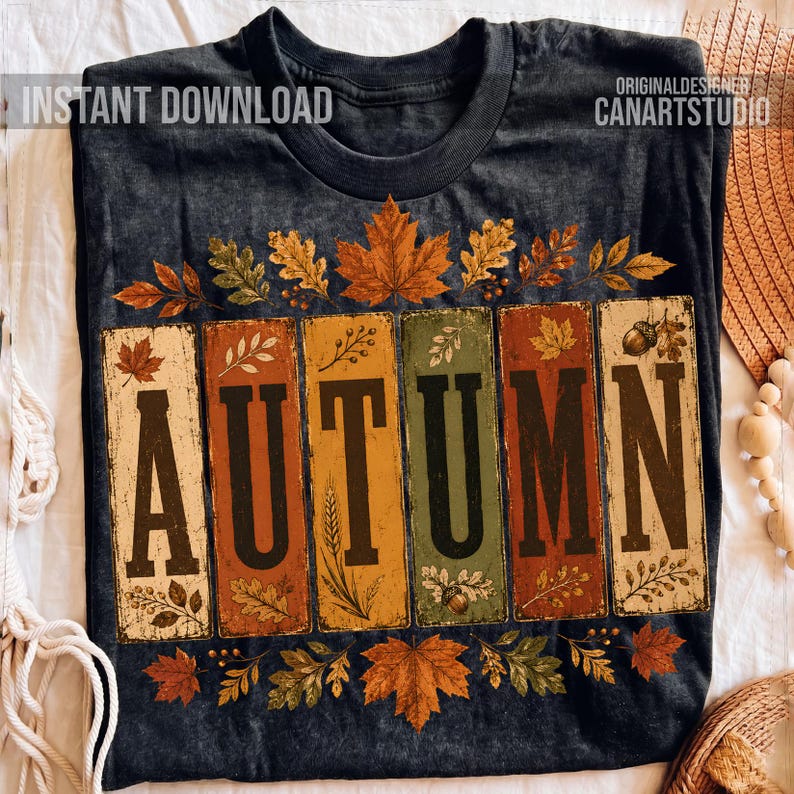 May include: A dark gray crew neck t-shirt featuring the word AUTUMN in large black letters. Each letter is set against a vertical rectangular panel in shades of orange, yellow, and green, decorated with illustrations of fall leaves and acorns.