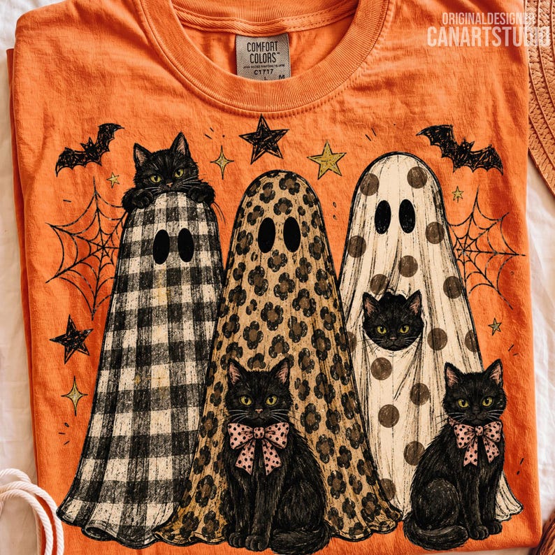 May include: An orange t-shirt featuring a graphic of three ghosts and three black cats. The ghosts have buffalo plaid, leopard print, and polka dot patterns. The cats have green eyes, and two wear pink bows. Bats, stars, and spiderwebs surround them.