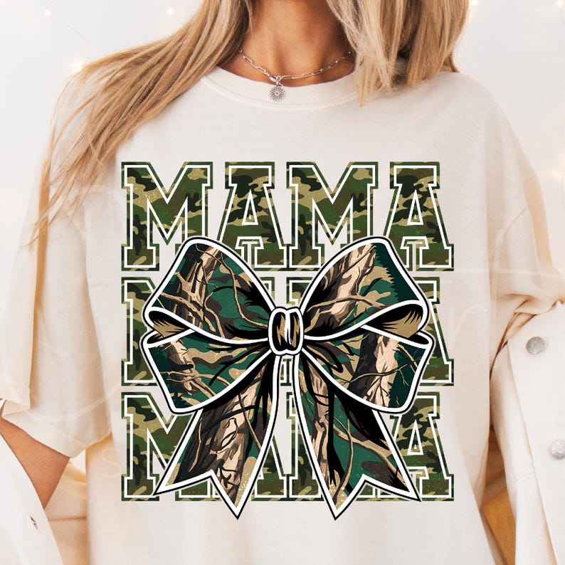 以下が含まれることがあります： 白地に緑と茶色の迷彩柄で「MAMA」とプリントされたTシャツ。大きな迷彩柄のリボンがTシャツの中央に配置されています。
