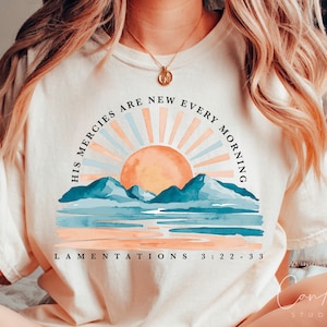 Op de afbeelding: Een wit T-shirt met een zonsopgang-afbeelding en de tekst "His mercies are new every morning" en "Lamentations 3:22-33".