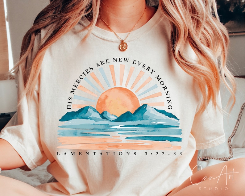 Op de afbeelding: Een wit T-shirt met een zonsopgang-afbeelding en de tekst "His mercies are new every morning" en "Lamentations 3:22-33".