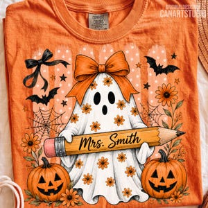 Puede incluir: Una camiseta naranja con una ilustraci&oacute;n de un fantasma blanco con flores naranjas, un lazo naranja grande y un l&aacute;piz con el texto Mrs. Smith. El dise&ntilde;o incluye murci&eacute;lagos negros, telara&ntilde;as y dos calabazas con flores naranjas.