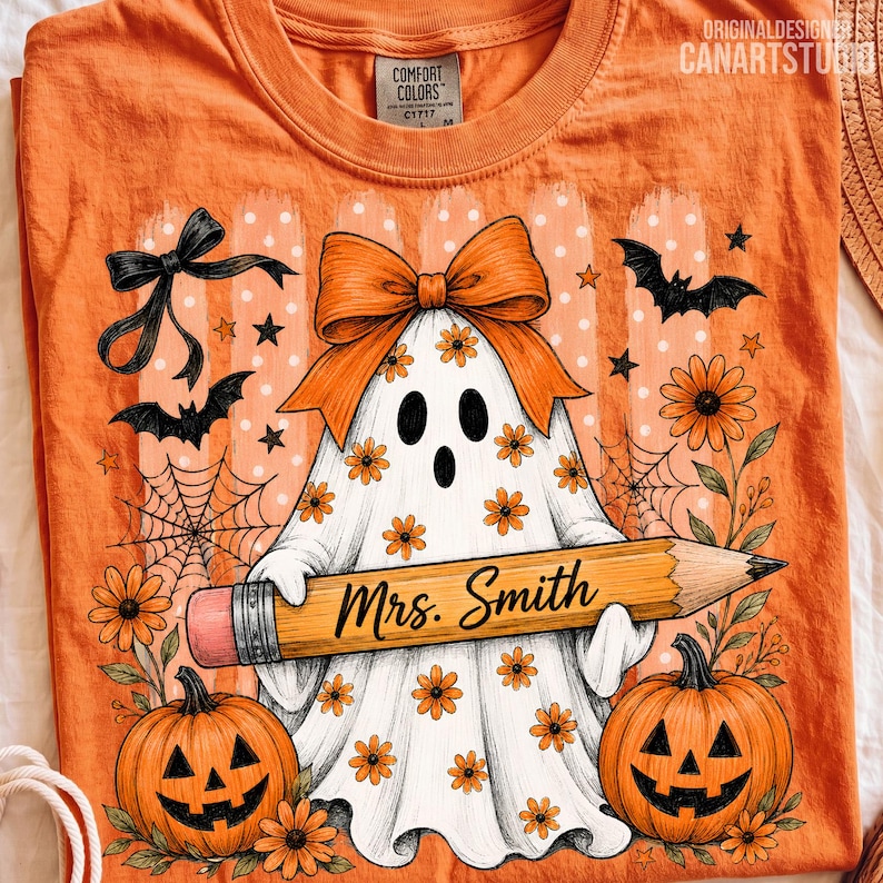 Puede incluir: Una camiseta naranja con una ilustraci&oacute;n de un fantasma blanco con flores naranjas, un lazo naranja grande y un l&aacute;piz con el texto Mrs. Smith. El dise&ntilde;o incluye murci&eacute;lagos negros, telara&ntilde;as y dos calabazas con flores naranjas.