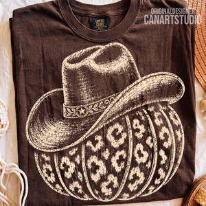 Può includere: Una t-shirt marrone scuro con una grafica color crema raffigurante un cappello da cowboy sopra una zucca con stampa leopardata. Il testo ORIGINALDESIGNER CANARTSTUDIO è stampato nell'angolo in alto a destra del tessuto della maglietta.