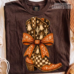 May include: A dark brown t-shirt featuring a graphic of a cowboy boot decorated with a large orange floral bow. The boot pattern includes white ghost figures wearing cowboy hats, cacti, and stars, with a checkered design on the lower section.