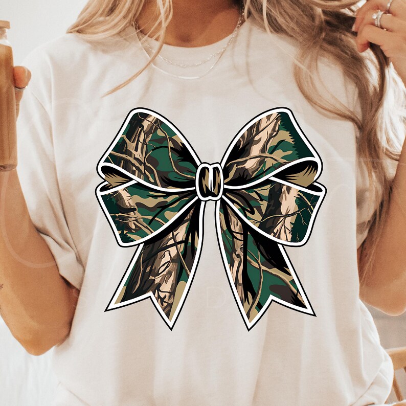 以下が含まれることがあります： 緑、茶色、白の迷彩柄のリボンがついた白いTシャツ