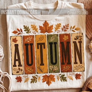 May include: A cream-colored t-shirt featuring the word AUTUMN in large black letters, each letter set within a vertical rectangular panel of varying fall colors. The design includes illustrations of maple leaves, oak leaves, and acorns.