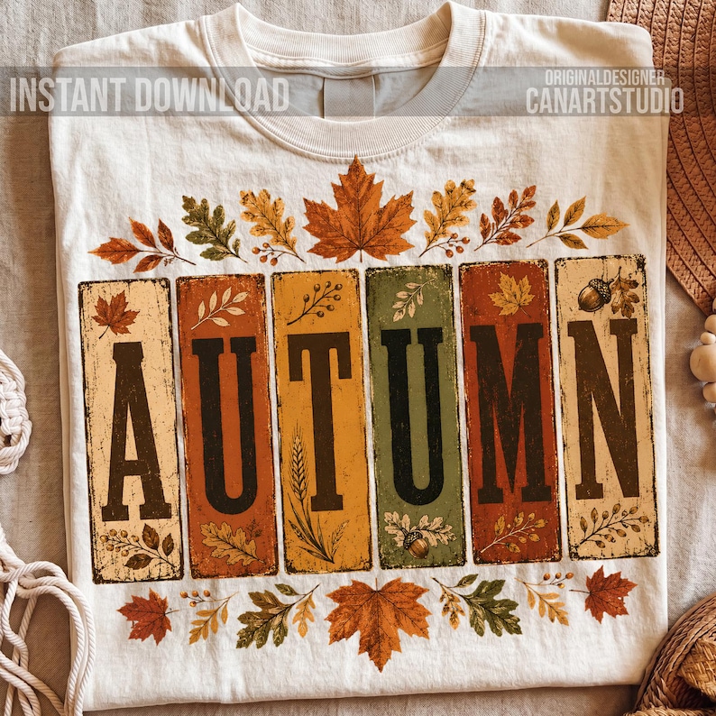 May include: A cream-colored t-shirt featuring the word AUTUMN in large black letters, each letter set within a vertical rectangular panel of varying fall colors. The design includes illustrations of maple leaves, oak leaves, and acorns.