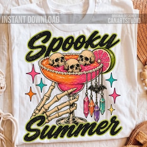 May include: A white t-shirt featuring a graphic of a skeleton hand holding a margarita glass filled with pink liquid and three skulls. The design includes a lime slice, hanging crystal charms, and the text "Spooky Summer" in black with green outlines.