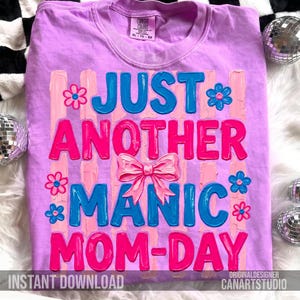 Op de afbeelding: Een lila T-shirt met de tekst "JUST ANOTHER MANIC MOM-DAY" in blauw en roze. Het ontwerp bevat een roze strik en bloemaccenten. Het shirt heeft ook de tekst "INSTANT DOWNLOAD" en "ORIGINALDESIGNER CANARTSTUDIO".
