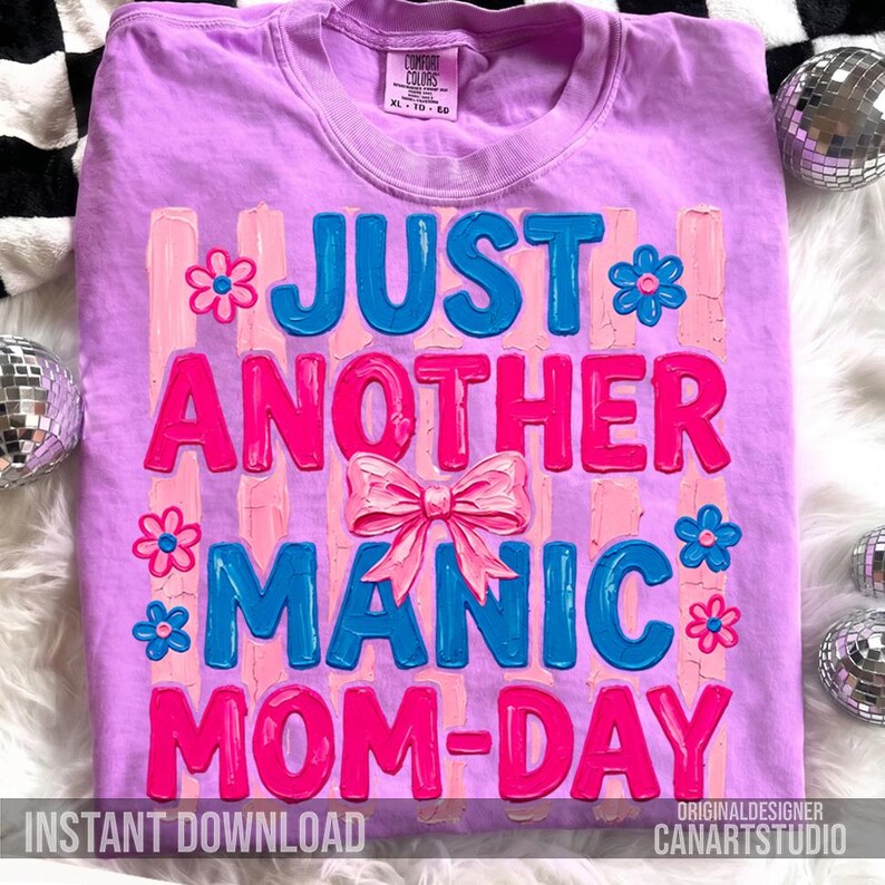 Op de afbeelding: Een lila T-shirt met de tekst "JUST ANOTHER MANIC MOM-DAY" in blauw en roze. Het ontwerp bevat een roze strik en bloemaccenten. Het shirt heeft ook de tekst "INSTANT DOWNLOAD" en "ORIGINALDESIGNER CANARTSTUDIO".