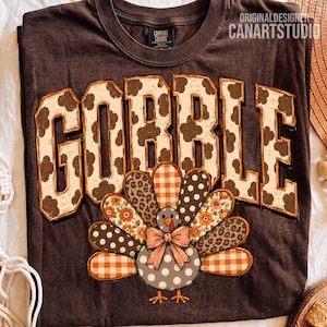 May include: A dark brown t-shirt featuring the word GOBBLE in large, blocky, cream-colored letters with a brown cow-print pattern. Below the text is a graphic of a turkey with a patterned tail featuring orange gingham, leopard print, and floral designs.