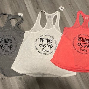 May include: Three tank tops with a circle design that says "Great Memories, Great Laughs, Great Times, Great Friends, Birthday Trip 2022". The tank tops are gray, white, and pink.