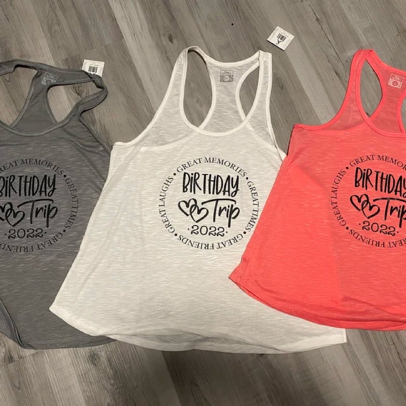 May include: Three tank tops with a circle design that says "Great Memories, Great Laughs, Great Times, Great Friends, Birthday Trip 2022". The tank tops are gray, white, and pink.