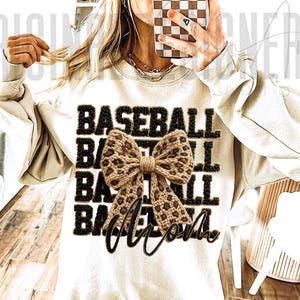 May include: Cream-colored sweatshirt with the word "BASEBALL" in black, outlined in a textured, dark brown material. A leopard print bow is centered on the shirt, with the word "Mom" in a cursive font below the bow. The sweatshirt has long sleeves.