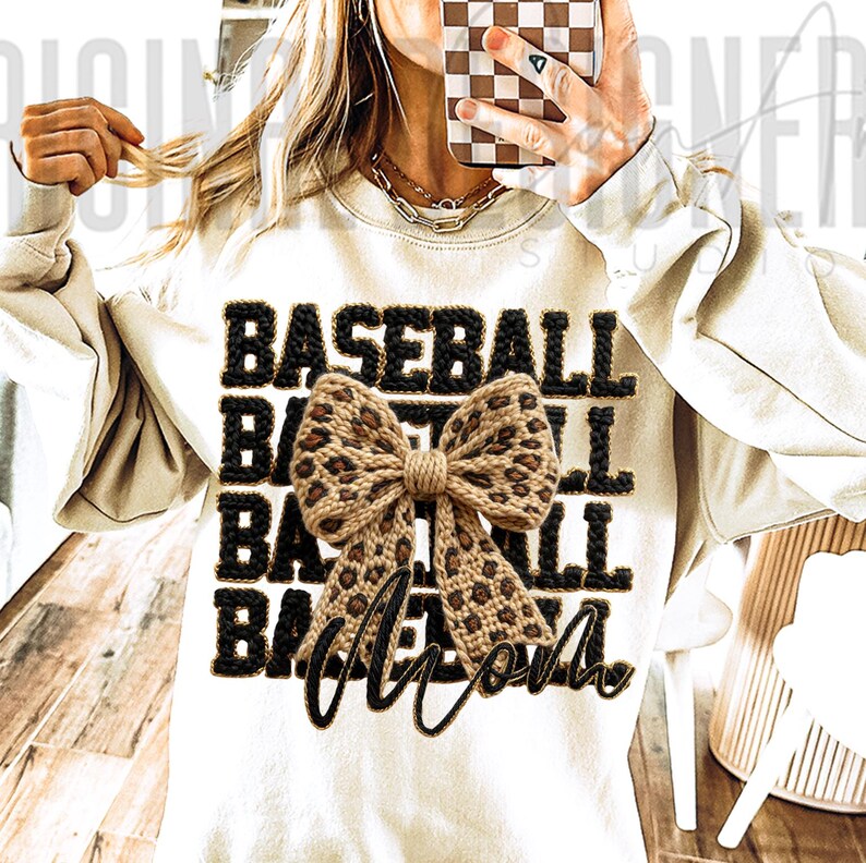 May include: Cream-colored sweatshirt with the word "BASEBALL" in black, outlined in a textured, dark brown material. A leopard print bow is centered on the shirt, with the word "Mom" in a cursive font below the bow. The sweatshirt has long sleeves.