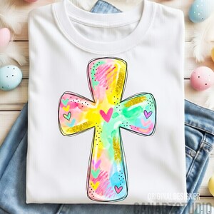 Op de afbeelding: Wit T-shirt met een kleurrijk kruisontwerp met een aquareleffect. Het kruis heeft gouden glitteraccenten en kleine hartdetails. Het shirt wordt gepresenteerd op een blauwe jeans, met paaseieren op de achtergrond.