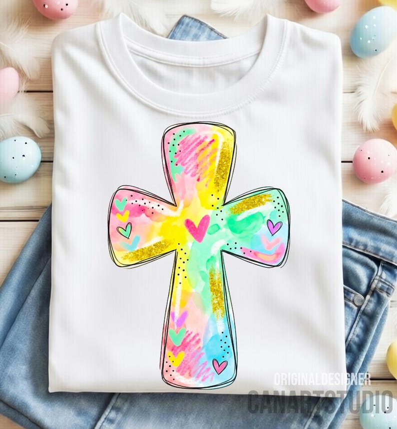 Op de afbeelding: Wit T-shirt met een kleurrijk kruisontwerp met een aquareleffect. Het kruis heeft gouden glitteraccenten en kleine hartdetails. Het shirt wordt gepresenteerd op een blauwe jeans, met paaseieren op de achtergrond.