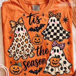 May include: An orange t-shirt featuring the text 'tis the season' in black script. The design includes three ghosts with pink cheeks, wearing bows and patterns of leopard print, black and white checkers, and black polka dots, alongside pumpkins and bats.