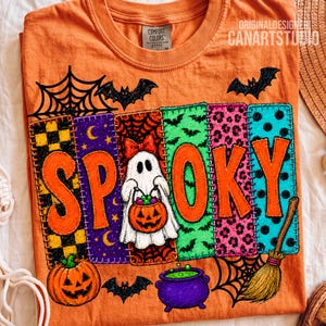 May include: An orange t-shirt featuring a graphic with the text SPOOKY. The design includes a white ghost holding a jack-o'-lantern, a purple cauldron, a broom, bats, and spiderwebs. The letters are set against colorful patterned panels.