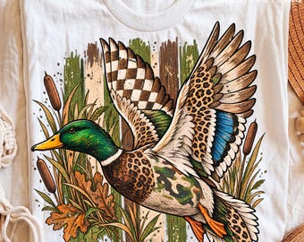 May include: A white t-shirt featuring a graphic of a mallard duck in flight. The duck has a green head, a yellow beak, and wings patterned with leopard print, camouflage, and a checkered design. The background shows cattails and autumn leaves.