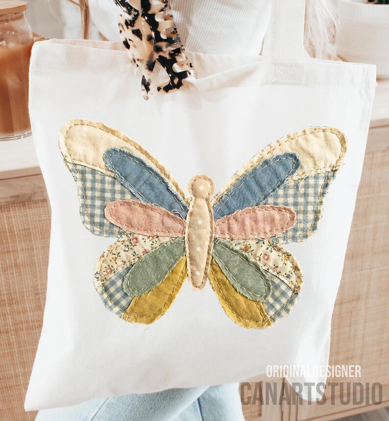 May include: White tote bag featuring a colorful butterfly applique. The butterfly design incorporates various fabrics in shades of blue, pink, yellow, and green. The text "ORIGINAL DESIGNER CANARTSTUDIO" is printed on the bottom right corner.