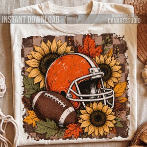May include: A cream-colored t-shirt featuring a graphic print of an orange football helmet, a brown football, yellow sunflowers, and orange autumn leaves set against a brown and cream checkered background with the text INSTANT DOWNLOAD CANARTSTUDIO.