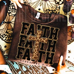 May include: A brown sweatshirt with the word "FAITH" in a stacked design, with a cross in the center. The text "EPHESIANS 2:8" is below the word "FAITH". The letters and cross are in black and tan.