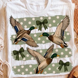 Pu&ograve; includere: Una t-shirt bianca con una stampa grafica di tre germani reali in volo su uno sfondo verde salvia a pois bianchi. Il design include tre fiocchi verde scuro intervallati tra le anatre, con il testo ORIGINALDESIGN CANARTSTUDIO.
