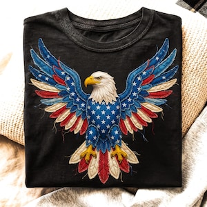 May include: Black t-shirt featuring a detailed graphic of an American bald eagle with outstretched wings. The eagle's wings are designed with the colors of the American flag: blue with white stars, and red and white stripes. The eagle has a white head and yellow beak.