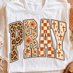 Könnte beinhalten: Ein weißes T-Shirt mit dem Wort PRAY in großen Blockbuchstaben. Das P hat ein Leopardenmuster, das R zeigt orangefarbene Blumen und grüne Blätter, das A hat ein orange-weißes Karomuster und das Y zeigt orangefarbene Kreuze.