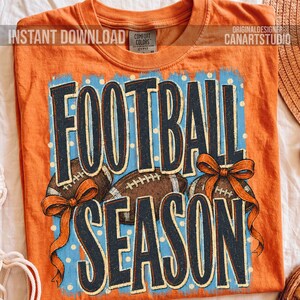 K&ouml;nnte beinhalten: Ein orangefarbenes T-Shirt mit kurzen &Auml;rmeln und einem Grafikdruck mit dem Text FOOTBALL SEASON in dunkelblauen Buchstaben. Das Design zeigt zwei braune Footballs und zwei orangefarbene Schleifen auf hellblauem Grund mit wei&szlig;en Punkten.