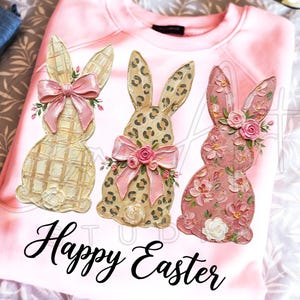 Puede incluir: Sudadera rosa claro con tres dise&ntilde;os decorativos de conejos de Pascua. Los conejos tienen dise&ntilde;os a cuadros, estampado de leopardo y floral. Cada conejo tiene un lazo rosa y adornos florales. Las palabras "Happy Easter" est&aacute;n escritas en cursiva.