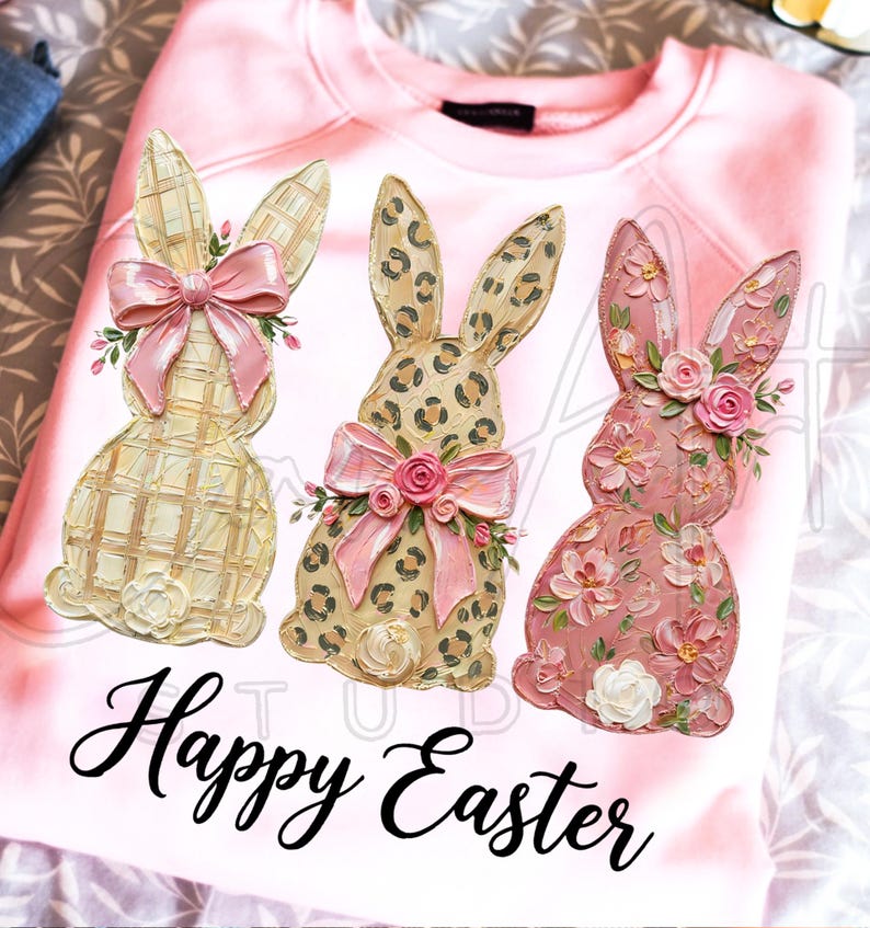 Puede incluir: Sudadera rosa claro con tres dise&ntilde;os decorativos de conejos de Pascua. Los conejos tienen dise&ntilde;os a cuadros, estampado de leopardo y floral. Cada conejo tiene un lazo rosa y adornos florales. Las palabras "Happy Easter" est&aacute;n escritas en cursiva.