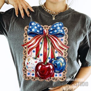 May include: Dark gray t-shirt featuring a patriotic design with a red, white, and blue bow and cherries. The bow has a blue star pattern, and the cherries are red, white, and blue. The shirt has the text "ORIGINAL DESIGNER ART STUDIO".