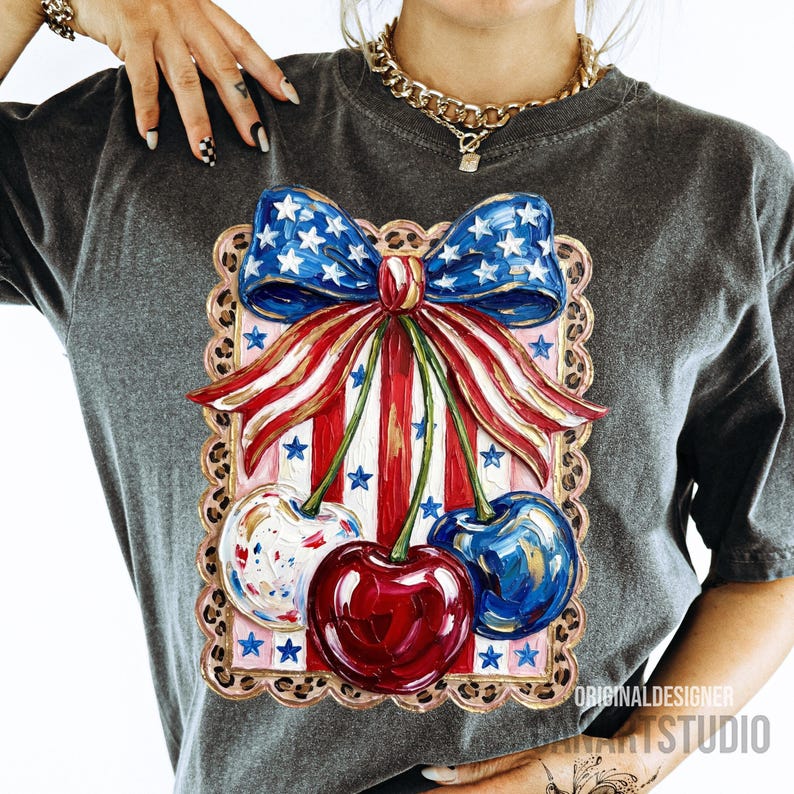 May include: Dark gray t-shirt featuring a patriotic design with a red, white, and blue bow and cherries. The bow has a blue star pattern, and the cherries are red, white, and blue. The shirt has the text "ORIGINAL DESIGNER ART STUDIO".